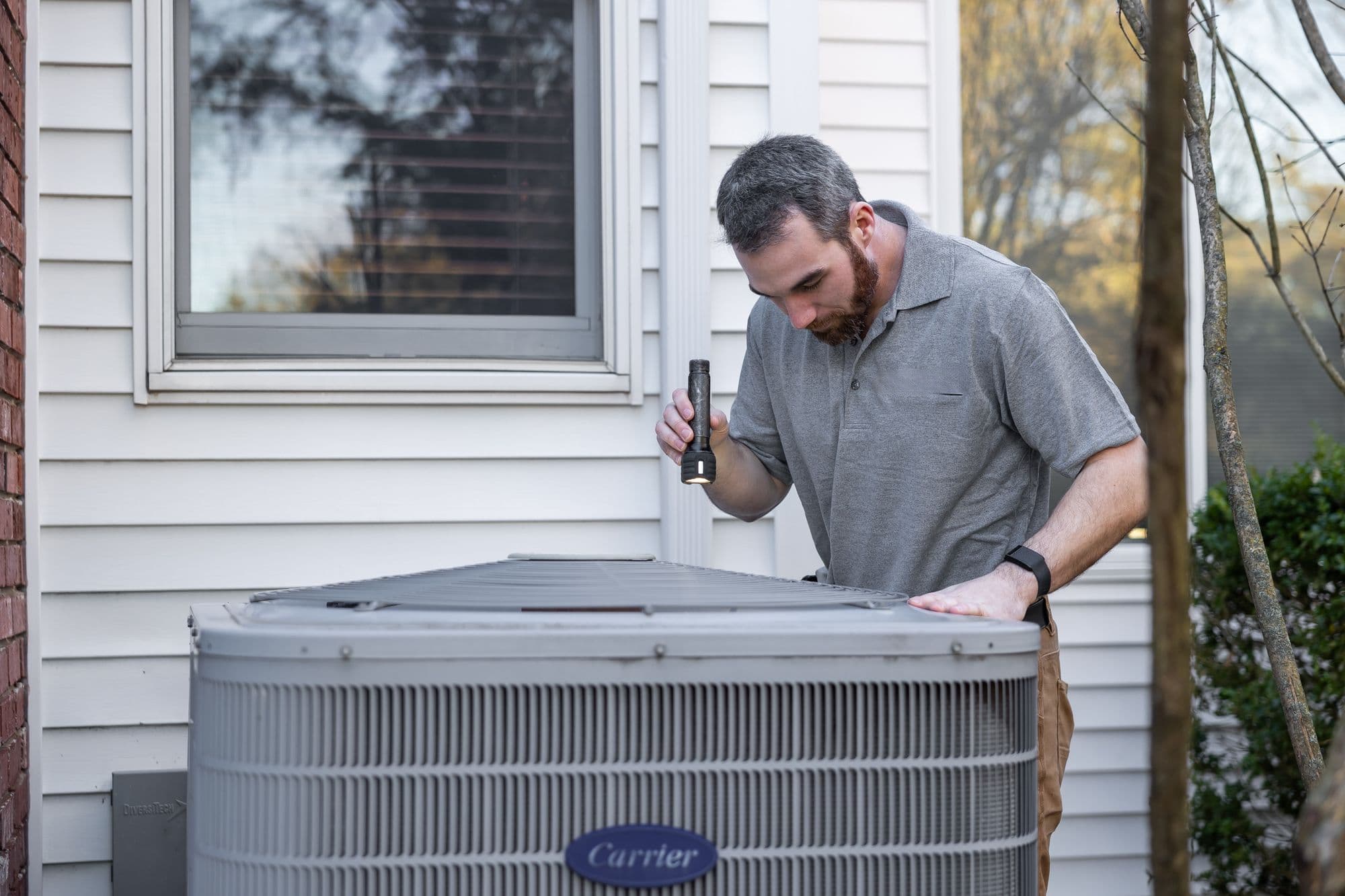 An HVAC technician shines a flashlight while inspecting an outdoor residential air conditioner condenser beside a house.